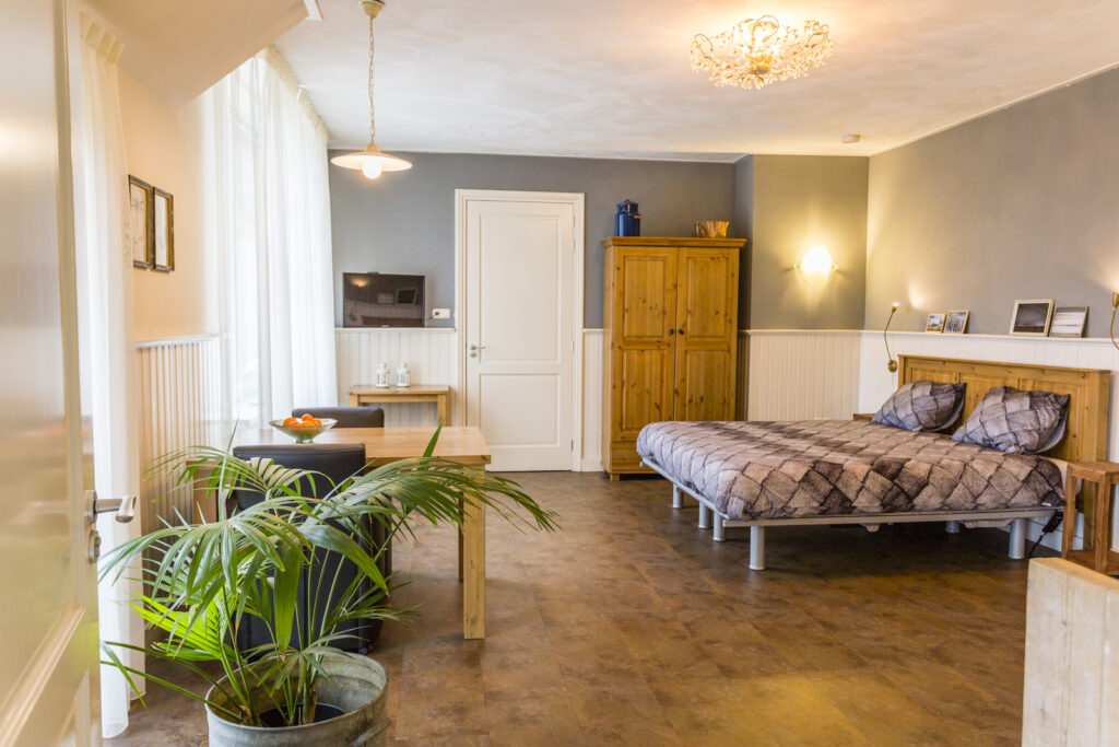 Waddenkamer, veel ruimte rondom het bed. Houtkleurige vloer met witte lambrisering en blauw erboven. Eettafeltje hout en tweepersoonsbed