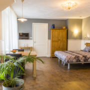 Waddenkamer, veel ruimte rondom het bed. Houtkleurige vloer met witte lambrisering en blauw erboven. Eettafeltje hout en tweepersoonsbed