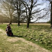 Eelke in haar elektrische rolstoel op een wat hobbelige graspad (holtingerveld) lang een rij bomen met weids uitzicht